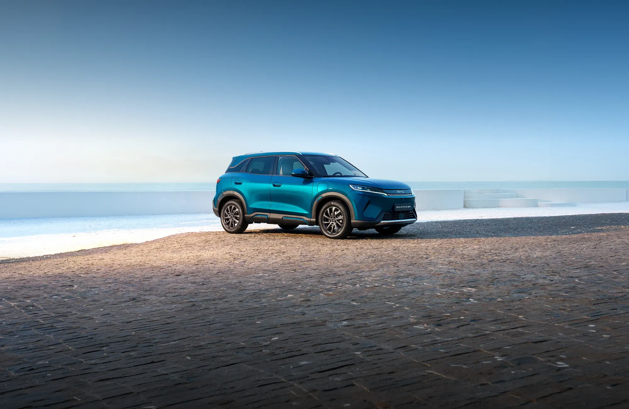 Um BYD ATTO 2 DM-i de cor azul-turquesa metalizado, estacionado de perfil numa praia arenosa com o mar e um céu azul limpo em fundo.