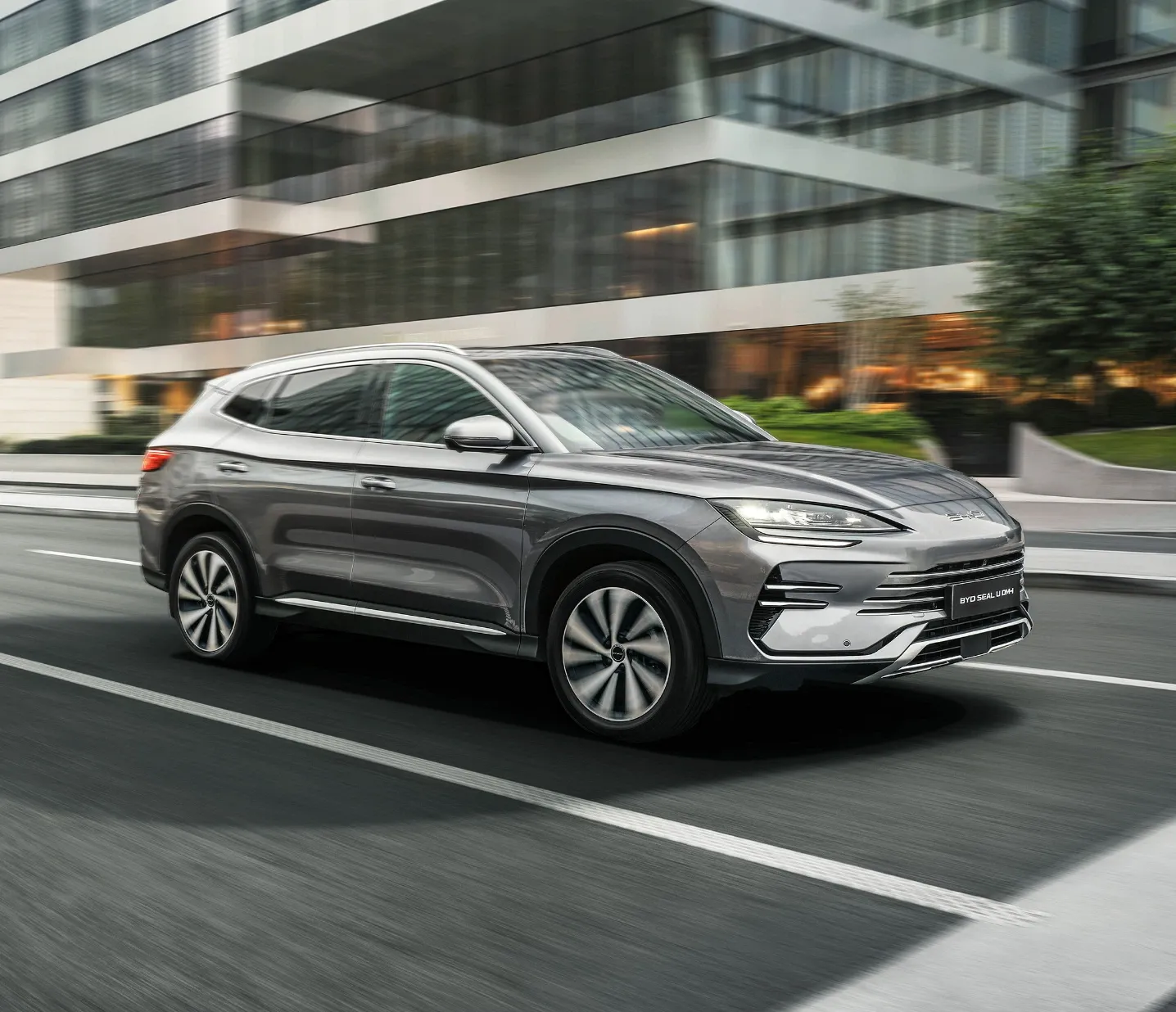 SUV BYD Seal U DM-i cinzento-escuro em movimento numa estrada urbana com edifícios modernos ao fundo.