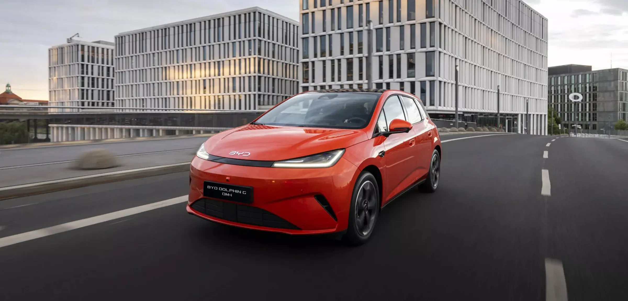 Um BYD Dolphin laranja vibrante a circular numa estrada de asfalto numa cidade, com arquitetura de escritórios moderna em vidro em segundo plano.