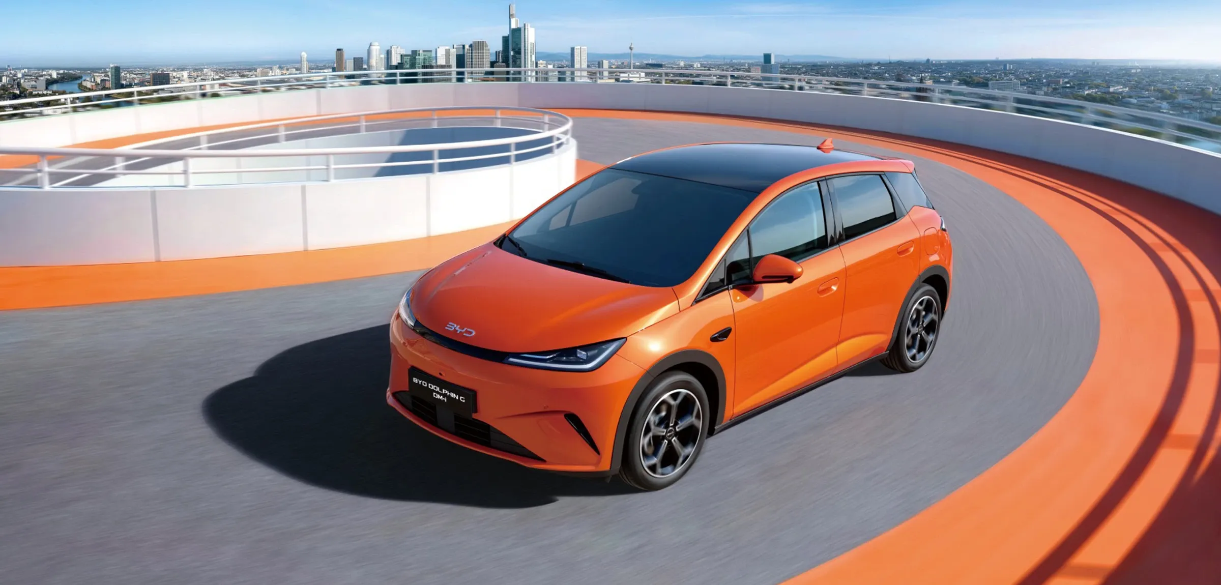 BYD DOLPHIN G DM-i, um hatchback moderno na cor laranja vibrante, a circular numa rampa em espiral com o horizonte de uma cidade ao fundo.