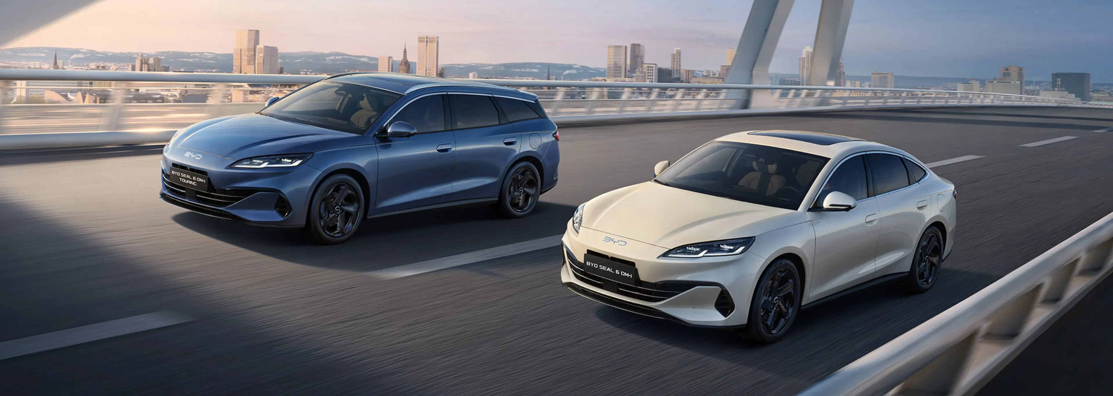"Dois carros BYD elétricos, um SUV azul e um sedã branco, estão lado a lado em uma estrada movimentada, com uma paisagem urbana moderna ao fundo."