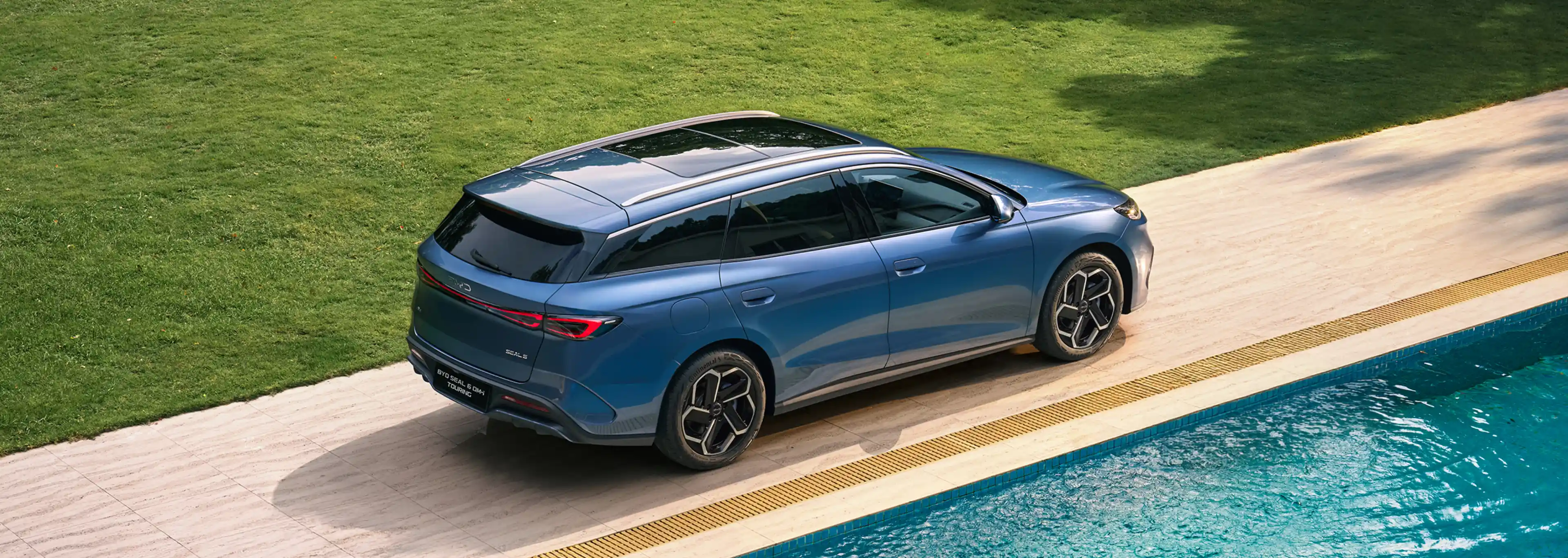 "Vista superior traseira de um BYD SEAL 6 DM-i Touring azul estacionado junto a uma piscina e relvado."