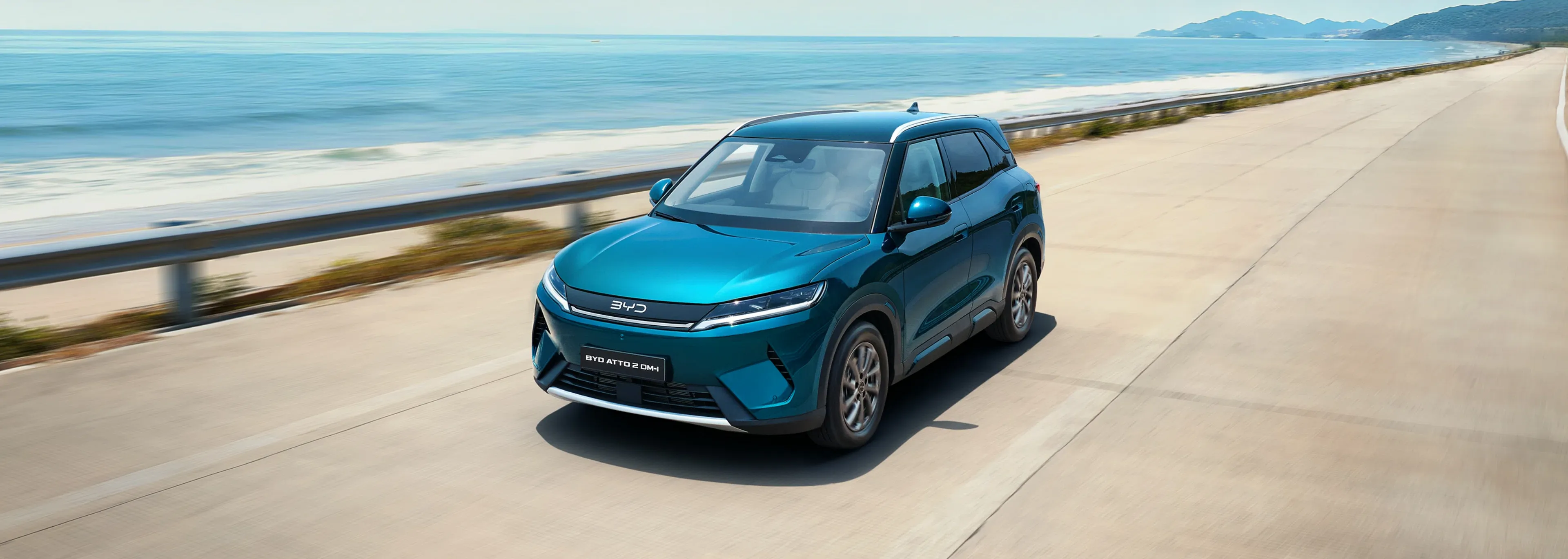 Um SUV BYD ATTO 2 DM-i de cor azul-turquesa a circular numa estrada à beira-mar.