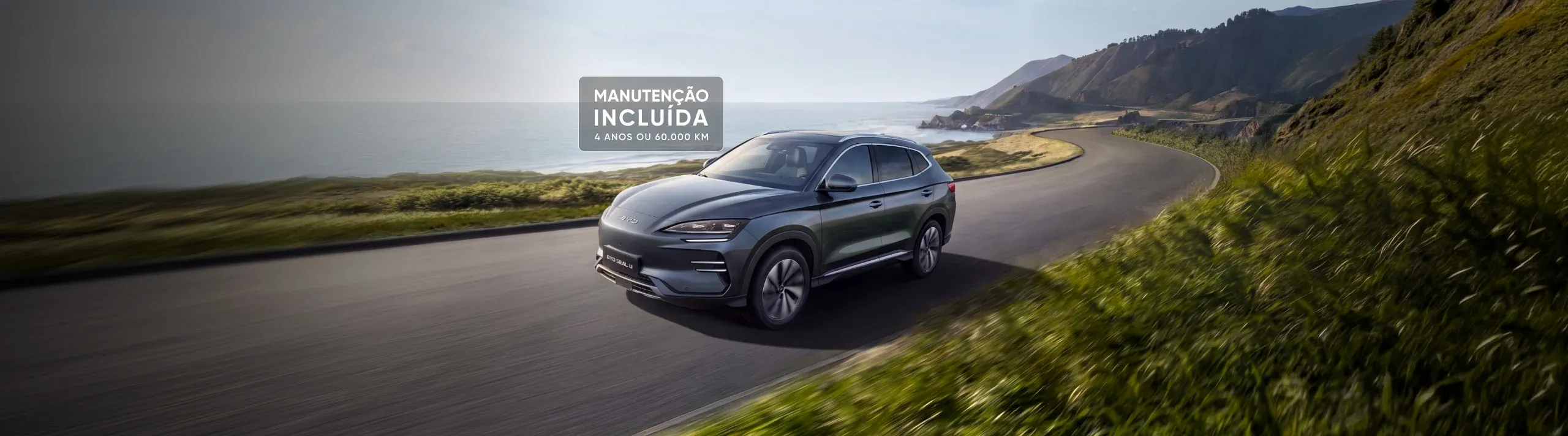 SUV BYD SEAL U cinzento a circular numa estrada costeira cénica, com um destaque visual onde se lê "MANUTENÇÃO INCLUÍDA: 4 ANOS OU 60.000 KM".