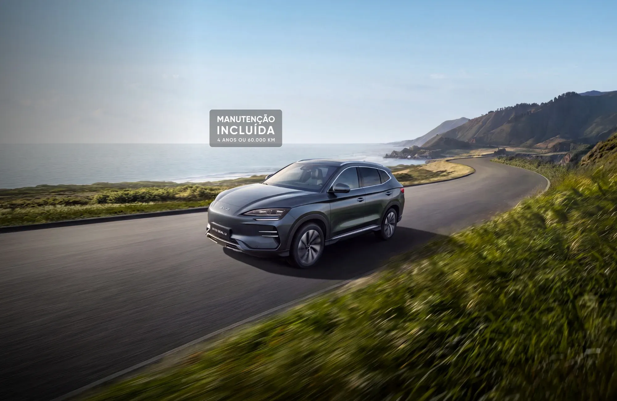 SUV BYD SEAL U cinzento a circular numa estrada costeira cénica, com um destaque visual onde se lê "MANUTENÇÃO INCLUÍDA: 4 ANOS OU 60.000 KM".