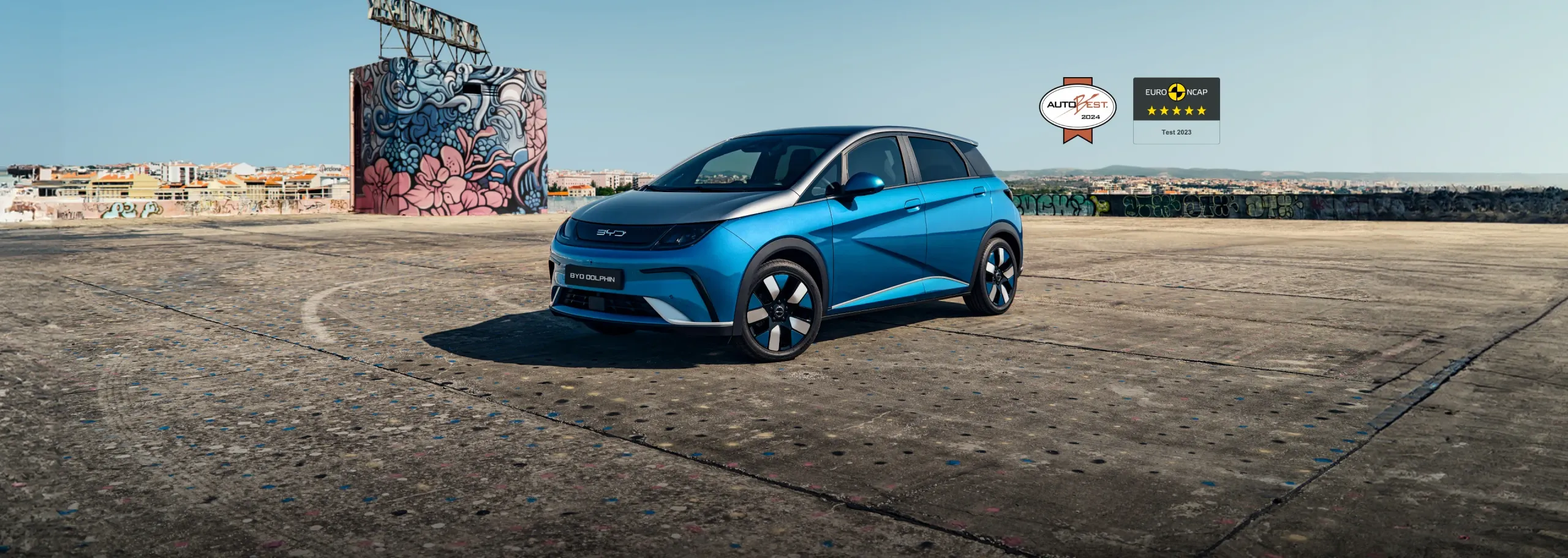 Vista a três quartos de um BYD DOLPHIN azul numa área de betão e com um bloco grafitado mais atrás