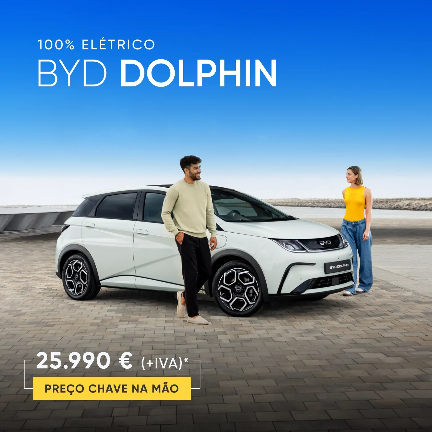 Um homem e uma mulher ao lado de um carro elétrico BYD DOLPHIN branco num espaço exterior com vista para o mar e céu azul.