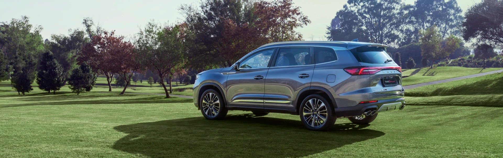 Chery Tiggo 7 cinza estacionado em relvado arborizado com iluminação natural de fim de tarde.