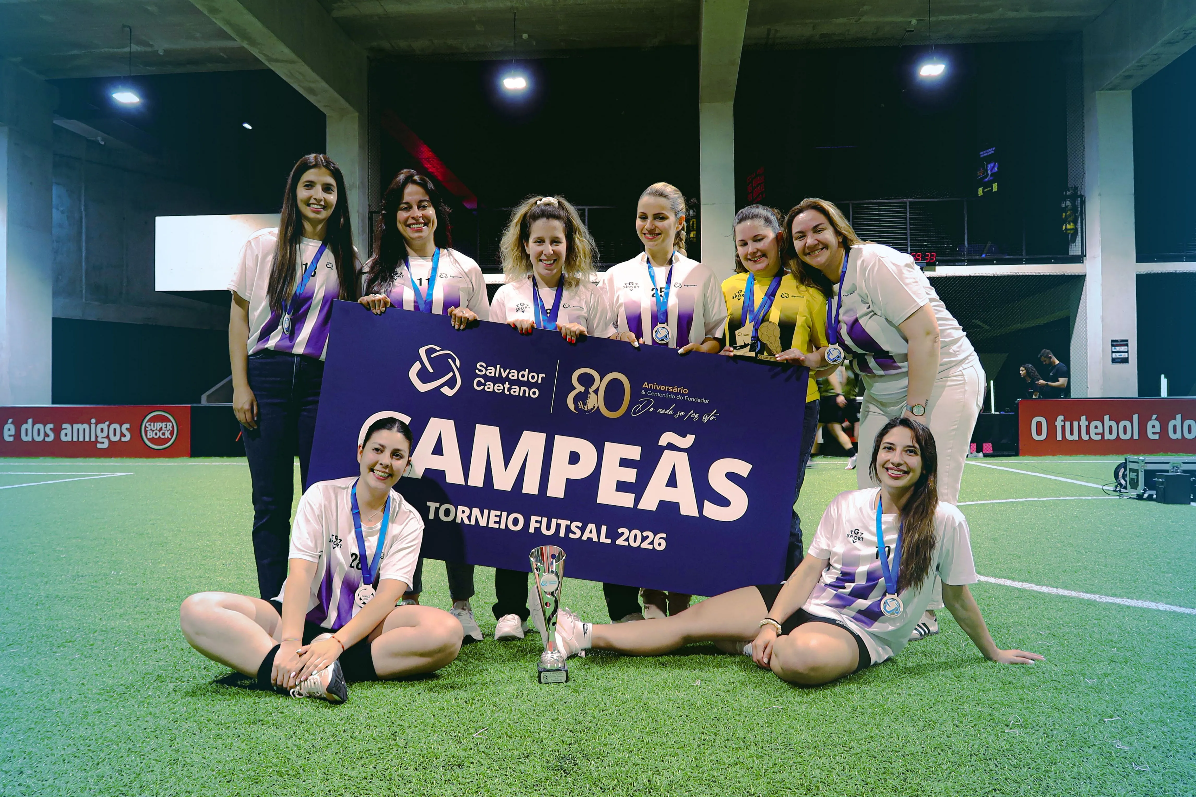 oito mulheres no relvado de um campo de futsal com cartaz de campeãs do torneio de futsal 2026 da Salvador Caetano