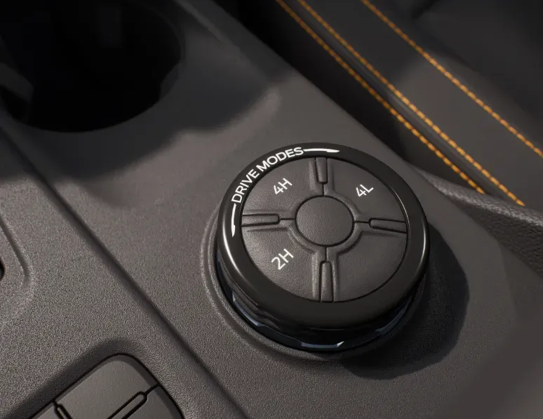 A close-up image of a car's center console showing a rotary dial with multiple drive mode options, set against a dark interior with orange stitching.