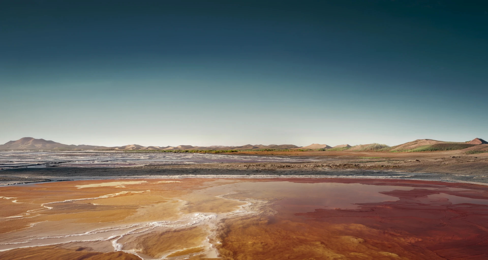 Paisagem natural árida com lago de água salgada em tons alaranjados e montanhas ao fundo sob um céu amplo.
